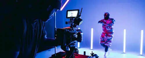 A camera operator films a person in colorful clothing dancing amid neon lights, with a monitor displaying the scene—perfect inspiration for your next shoot with studio hire Canberra.
