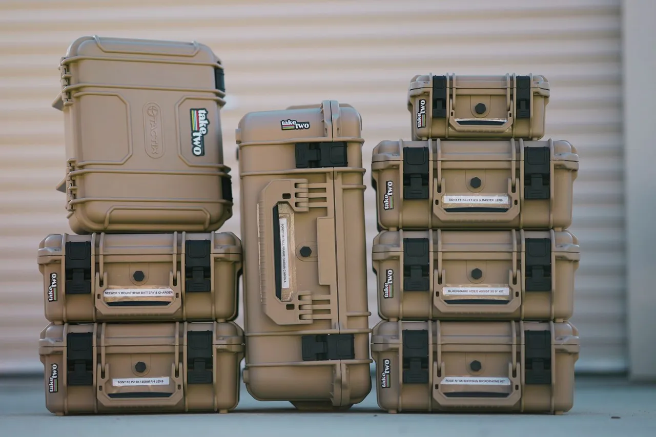 Stacked tan hard protective cases with black latches are arranged in three columns, standing on a concrete surface in front of a corrugated metal background.